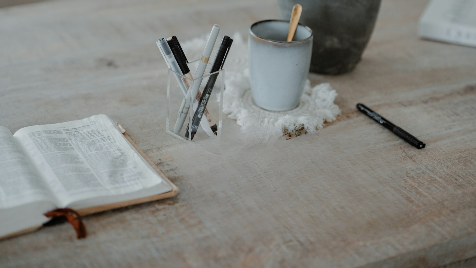 Open book, pen holder with pens, mug, and notebook on a wooden surface. For a blog post 15 scriptures to Bring into Your Everyday